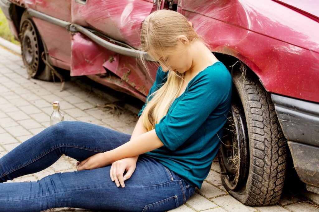 A teenage girl sitting down against a wrecked car.